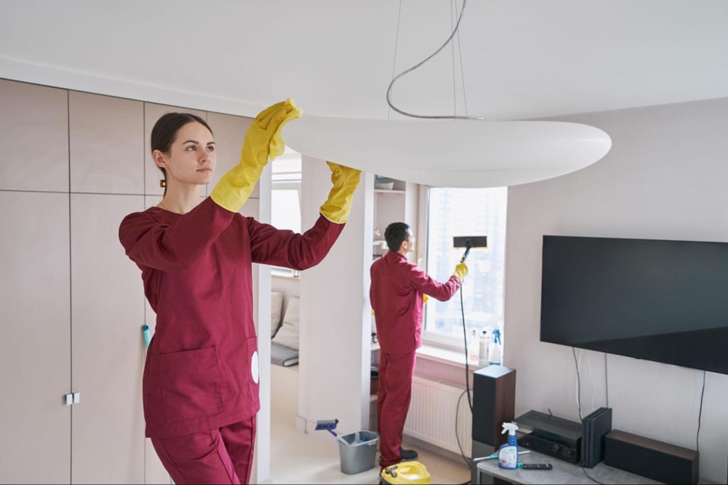 Two professional cleaners in maroon uniforms and yellow gloves perform eco-friendly deep cleaning in a living room.